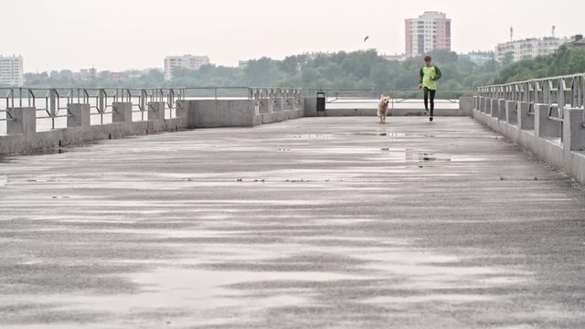 Boy running towards the camera along pier at cold autumn day with adorable playful retriever