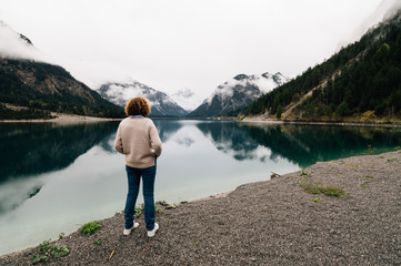 Fototapeta na wymiar Lonely woman looking at mountain lake with water reflections. Plansee lake located in Austria a cloudy and foggy day. Horizontal composition, space for copy