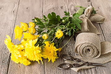 Bouquet of yellow chrysanthemums .
 Decorative tape of burlap, scissors and bouquet of yellow chrysanthemums on gray wooden table.