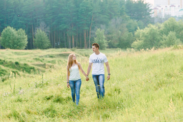 Naklejka na ściany i meble A lovely couple walking in a forest area, green grass all around them, holding hands,looking smiling at each other