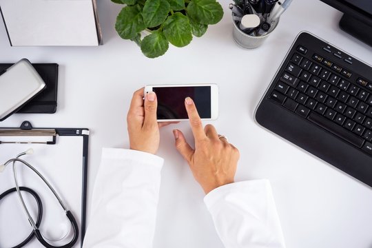 Female doctor touching black screen of a smart phone, on top of the working desk, inside her office.
