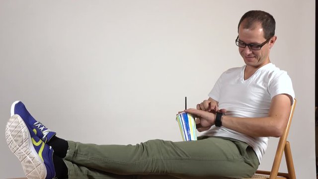 Man in black rim glasses holding plastic cup and using his smartwatch. Leisure time. Blanc space for adding your graphics or inscriptions. 4K shot