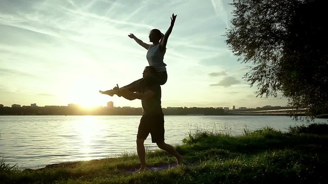 Acrobatic yoga on the bank of the river. Young man and woman practicing acrobatic yoga. The dark silhouettes of people in backlight. Wide angle. HD