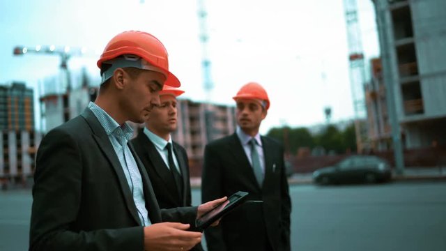 4k. UHD. Construction worker with digital pad and businessmen in suit and protective helmets talking on site about new construction. Crane and beams at the background. Teal and orange middle stabicam