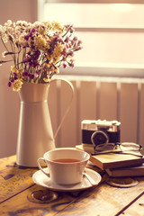 Taza de Té junto a libros, cámara de fotos y un florero con flores secas de colores sobre una mesa de madera junto a una ventana. Vista de frente