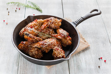 Chicken wings in cast-iron pan on wooden table. Marinated in tomato and honey sauce. Baked with sesame seeds.