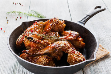 Chicken wings in cast-iron pan on wooden table. Marinated in tomato and honey sauce. Baked with sesame seeds.