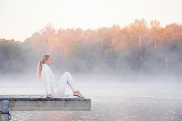 Woman wearing white clothes relaxing and practicing yoga in the mist on the lake footbridge early morning. The morning sun shines on the tree ends.