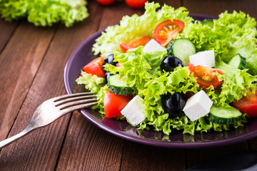 Greek salad (lettuce, tomatoes, feta cheese, cucumbers, black olives) on dark wooden background close up. Healthy food.