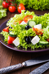 Greek salad (lettuce, tomatoes, feta cheese, cucumbers, black olives) on dark wooden background close up. Healthy food.