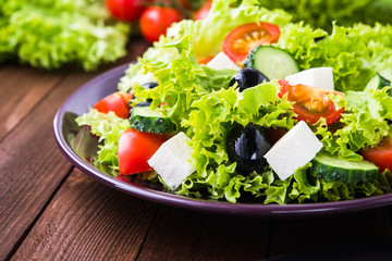 Greek salad (lettuce, tomatoes, feta cheese, cucumbers, black olives) on dark wooden background close up. Healthy food.
