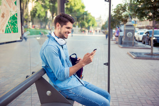 Young stylish man with headphones sitting on bus stop and typing