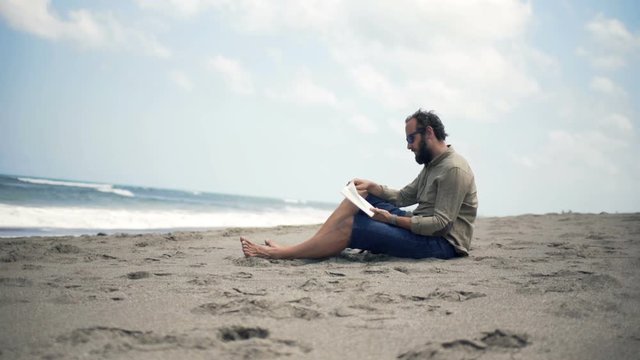 Young man reading book sitting on beach near sea

