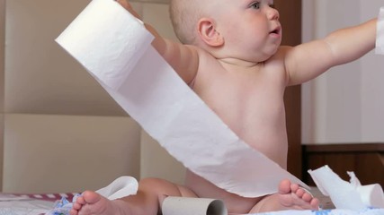 Attractive baby sitting on a bed and tearing toilet paper. Kid 1 year