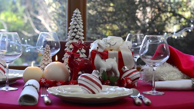 Stylish red and white Christmas table with reindeers and candles centerpiece in front of garden window, full table pan left.