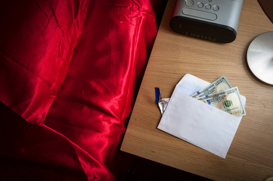 Prostitution concept with money in an envelope on a nightstand in a hotel room, representing the payment for a prostitute, escort or sex worker, next to the bed covered in red satin sheets