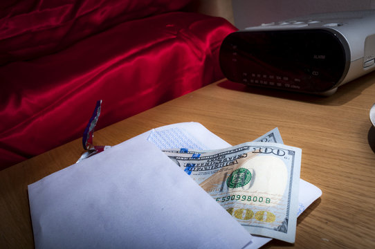 Prostitution concept with money in an envelope on a nightstand in a hotel room, representing the payment for a prostitute, escort or sex worker, next to the bed covered in red satin sheets