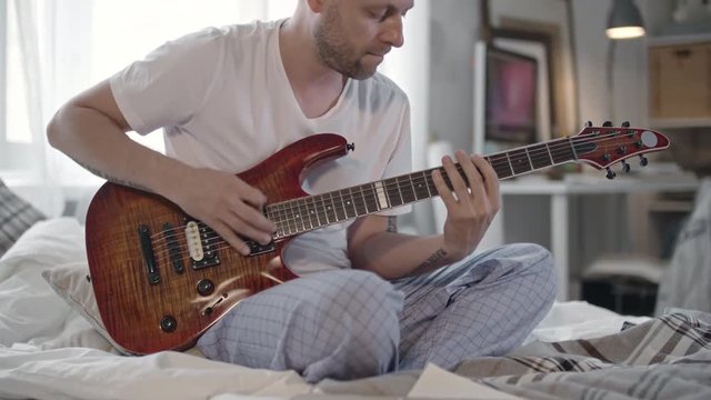 Man sitting on bed in pajamas and making some notes on paper while playing the guitar