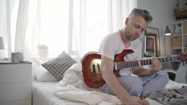 Man playing the guitar and writing down chords on paper while sitting on the bed