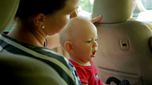 Mom goes with the kid in the back seat of the car. Cute little boy 1 year old goes to his mother's lap and looks around
