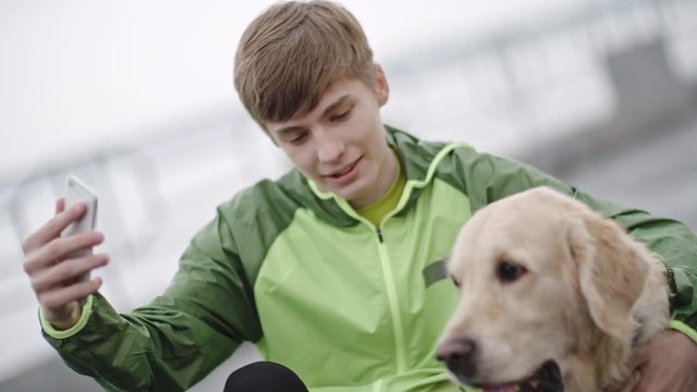 Medium shot of teenage boy hugging his golden retriever dog and taking selfie, then talking and petting him