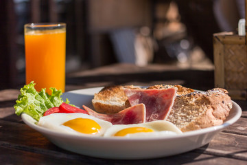 Breakfast of Ham, Bacon, Scrambled eggs, Tomato, Lettuce, Glass of Orange Juice and Toast close-up american breakfast on white ceramic plate on wooden table in Luxury Hotel Resort Spa on the mountain