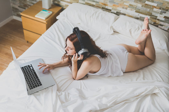 Red-haired woman in headphones using laptop on bed