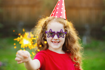Happy girl in party hat with burning sparkler in her hand.