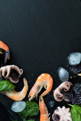 Zubereitung frischer Meeresfrüchte in der Küche mit rosa Gourmet-Garnelen und Tintenfischen, umgeben von Eis, frischen Kräutern und Gewürzen auf schwarzem Steinhintergrund