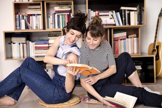 Two best friends sitting back to back on the floor of living room looking at a book