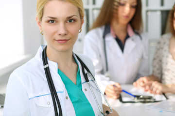 Young beautiful female doctor smiling  on the background with patient and his physician in hospital. High level and quality medical service concept.