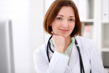 Young brunette female doctor sitting at a desk and working on the computer at the hospital office.  Health care, insurance and help concept. Physician ready to examine patient