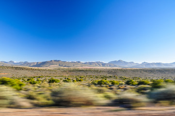 Driving on Route 62 road near Oudtshoorn with bushland and Swartberg mountain range in background.Western Cape province, The Karoo,South Africa.Oudtshoorn has many ostrich farms.Motion blur near road.