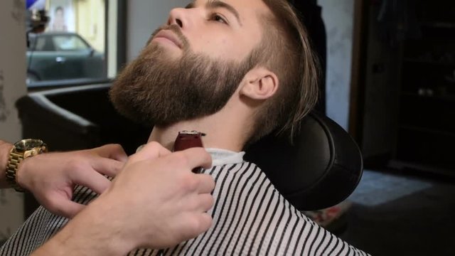 Hairdresser with tattoo serving client in barbershop, making beard haircut using shaver