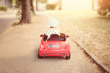  Little girl dressed in white blackheads shirt and hat, driving a red toy car near the park. Baby...