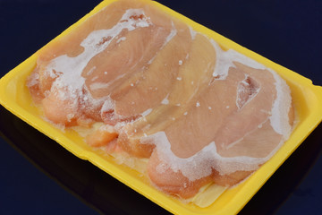 Frozen chicken breast tenderloin meat lightly covered with ice thawing in packaging on dark blue...