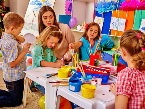 Children with teacher woman painting school on paper at table in kindergarten . Interior of kindergarten or primary school.