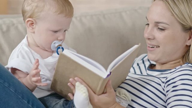 Toddler boy with pacifier sitting on his mother and looking at pictures in book