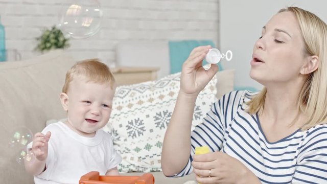 Blond mother blowing soap bubbles while her toddler son trying to catch them