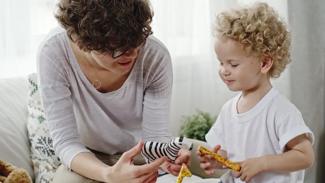 Curly mother in glasses and her blond toddler looking at wooden zebra toy and playing together