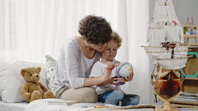 Curly mother in glasses sitting on bed with blond toddler boy and showing him desk globe