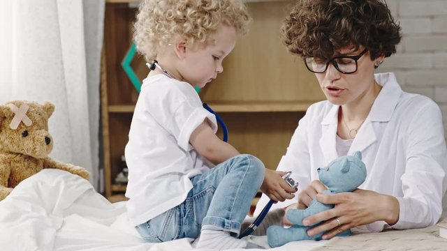 Toddler boy with curly blond hair holding stethoscope and caring for plush bear with mother
