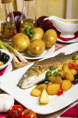 Fried sea bass with side dish on a plate, served on a table next to fresh vegetables and olive oil.