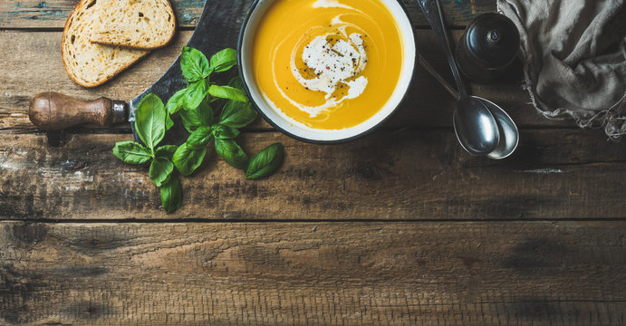 Pumpkin cream soup in bowl with fresh basil, spices and grilled bread slices over old rustic wooden background, top view, copy space, horizontal composition