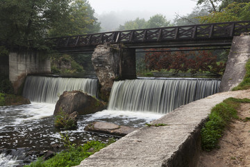Wooden bridge across the river with waterfall. Foggy morning.
