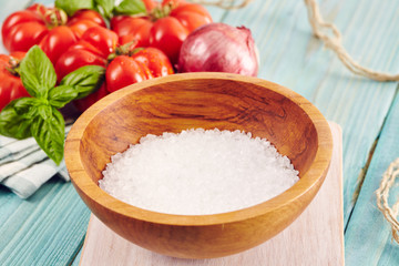 a wooden bowl with marine salt crystals surrounded by tomatoes and onion on an aquamarine wooden table