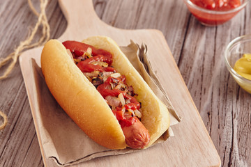 Hot dog with onion, ketchup and mustard on a white chopping board on a wooden table. Ketchup and mustard in two transparent bowls.