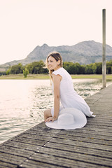 Portrait of a blonde woman sitting on a dock on a lake, with a white dress