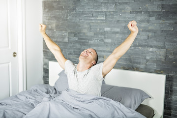 Cheerful young man waking up in bed and stretching his arms