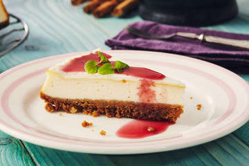 new york cheesecake with cherry sauce on a plate on a blue wooden table, full cake in background